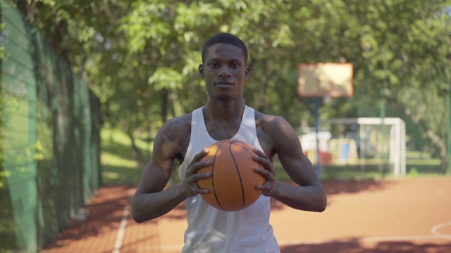 自信的非裔美国运动员在篮球场上拿着球摆姿势的肖像。一个严肃的年轻人站在户外看着摄像机。自信、运动、健康的生活方式。视频素材