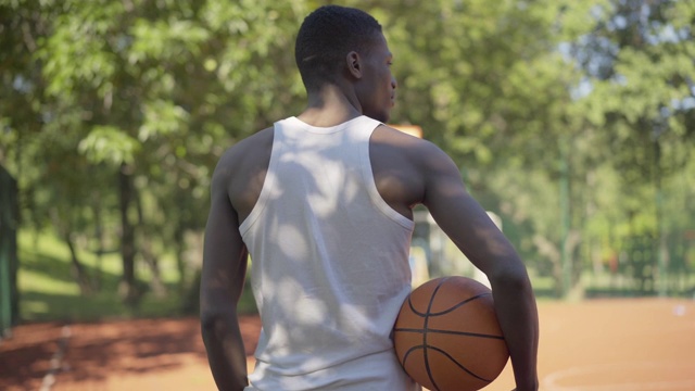 背景的非洲裔美国运动员站在阳光灿烂的篮球场上，白色的衬衫和球，转向镜头和微笑。年轻自信的英俊男子在户外摆姿势在阳光明媚的一天。视频素材