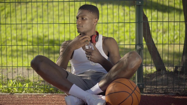 满意的篮球运动员坐在球场上，拿着球，喝着新鲜的冷水。在阳光明媚的一天，疲惫的年轻非洲裔美国人在比赛后休息。体育的概念。视频素材