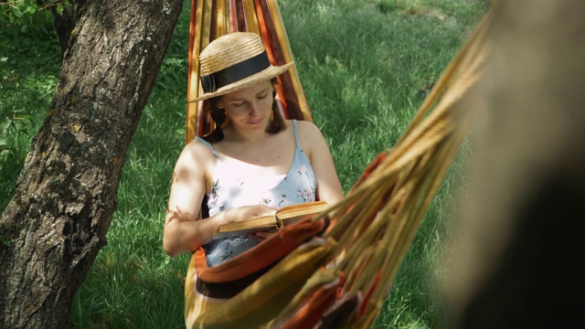 年轻的女士戴着帽子和书在吊床上。迷人的快乐女人躺在吊床上，在绿色花园读书。深色头发的女性躺在吊床上，手里拿着书，放松和享受假期视频素材