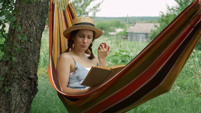 年轻的黑发女子戴着草帽躺在吊床上，一边看书一边吃苹果。快乐的黑发女性休息在吊床户外。在绿色花园的吊床上放松的女孩视频素材