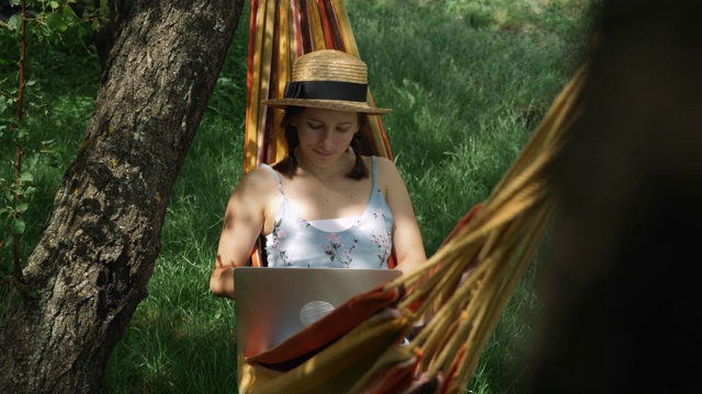 年轻的褐发女性戴着帽子躺在吊床上，在绿色花园的笔记本电脑上工作。迷人的女人躺在吊床上，在户外用笔记本电脑。暑假的概念视频素材