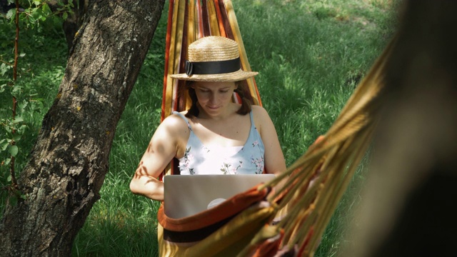 在吊床上用笔记本电脑的女人。戴着帽子的年轻女性自由职业者躺在吊床上，在户外用笔记本电脑工作。女孩在花园的吊床上休息，打开笔记本开始工作视频素材
