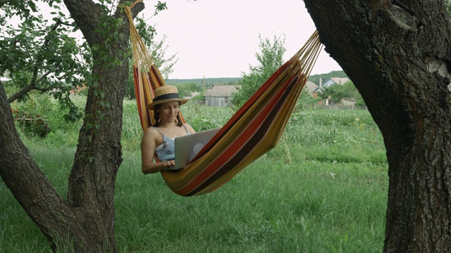 年轻的自由职业者躺在吊床上，手提电脑在户外。可爱迷人的女性穿着衣服和帽子坐在吊床上的自然和工作与笔记本电脑。女孩躺在吊床上用笔记本视频素材