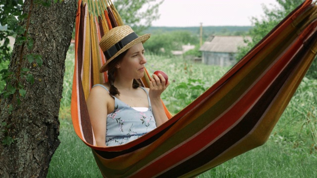 周末，快乐的女人在绿色花园的吊床上休息。年轻女子躺在吊床上，在乡下享受暑假。褐发女性，吃苹果，在吊床上休息视频素材