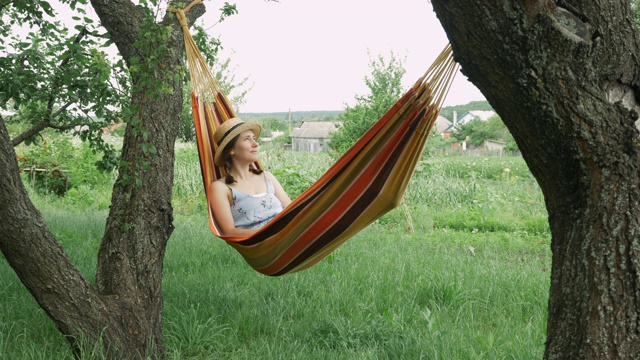 一个年轻女子躺在户外两棵树中间的吊床上休息。可爱的女孩躺在乡间的吊床上做白日梦。戴着帽子和穿着衣服的黑发女性在外面的吊床上放松视频素材