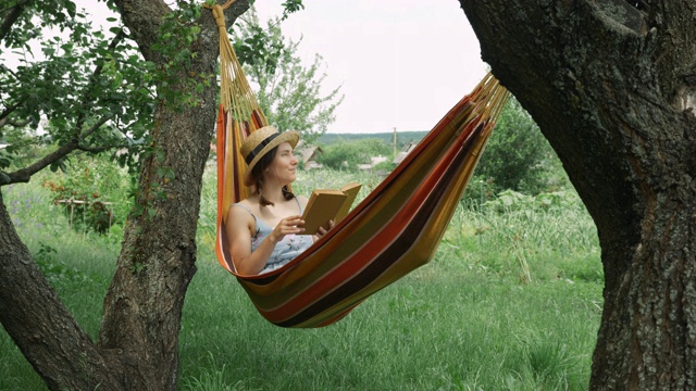 戴着帽子的年轻女子躺在绿色花园舒适的吊床上。可爱迷人的黑发女子在户外的吊床上看书。女孩躺在吊床上看书视频素材
