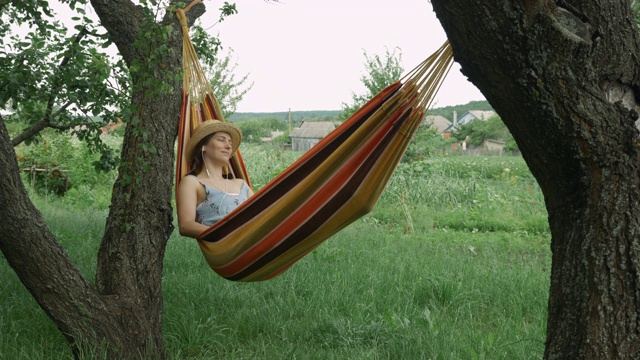 年轻女性戴着耳机在吊床上听音乐，休息和放松。一个戴着草帽的黑发女人躺在户外的吊床上听着耳机里的音乐视频素材