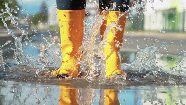 特写:穿着牛仔裤和黄色雨靴的女人跳在玻璃水坑里。视频下载