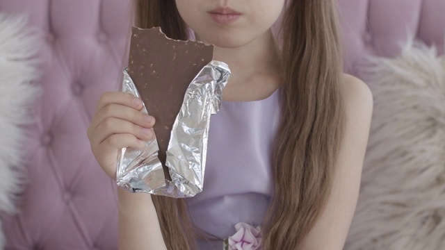 一个面目全非的白人小女孩在咬巧克力棒。可爱的深色头发的孩子在家里享受美味的甜点的特写。在室内吃不健康食品的孩子视频素材
