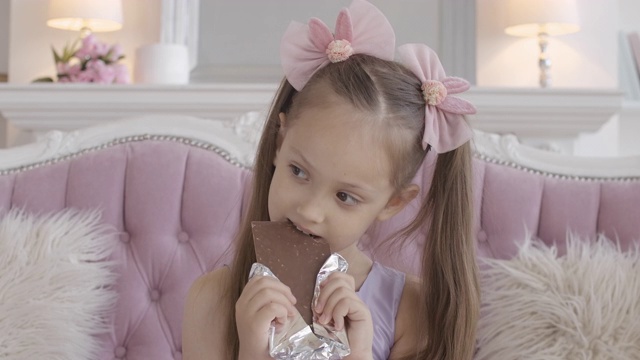 可爱的小女孩在吃巧克力。深色头发的白人小孩，一边嚼着甜食。在家享受不健康食物的孩子视频素材