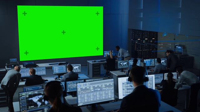 在一个控制中心的房间里，工程师和控制器在计算机上工作，大的绿色屏幕水平模拟。电信员工在布满显示器的监控室工作。视频下载