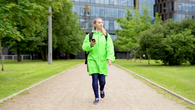 这是一名身穿绿色制服、带着保暖袋的十几岁女食品快递员的全身正面照片，她使用智能手机上的gps应用程序，在送货时四处查看视频下载