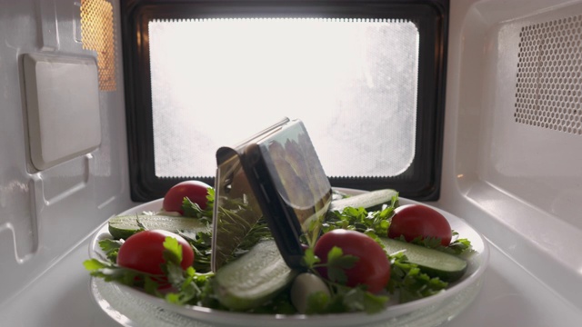 装有沙拉的盘子和微波炉屏幕上显示烹饪食物视频的手机。视频下载