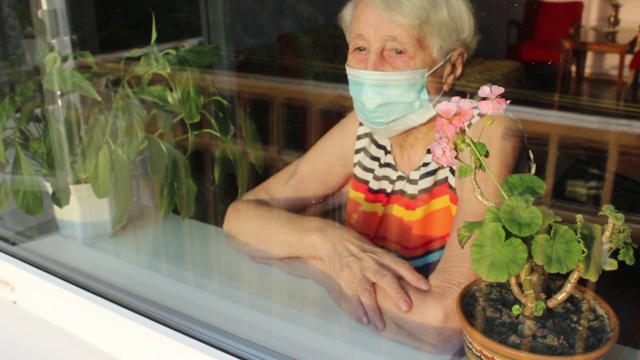 2019冠状病毒病、健康、安全和大流行概念——孤独的老年妇女戴着医用防护口罩，坐在家里靠近窗户的地方防止病毒感染视频下载
