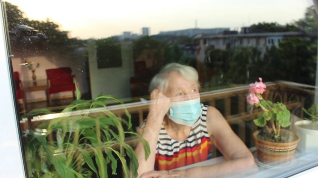 2019冠状病毒病、健康、安全和大流行概念——孤独的老年妇女戴着医用防护口罩，坐在家里靠近窗户的地方防止病毒感染视频素材
