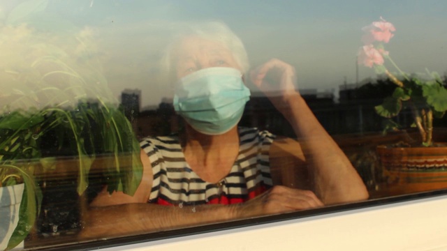 2019冠状病毒病、健康、安全和大流行概念——孤独的老年妇女戴着医用防护口罩，坐在家里靠近窗户的地方防止病毒感染视频素材