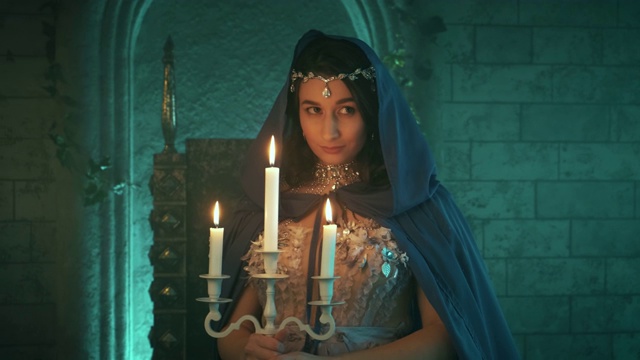 美丽的年轻神秘的童话故事的女人在万圣节服装中世纪女王摆模特在背景石墙城堡。黑发女孩拿着烛台，燃烧着蜡烛。蓝色创意斗篷，兜帽视频下载