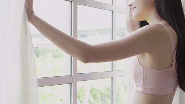 美丽年轻的亚洲女人醒来，走到窗户与开放的窗帘在早上舒适，性感的女孩在内衣微笑放松和休息在假期与快乐，生活方式的概念。视频下载