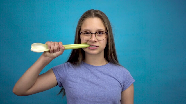 一个十几岁的女孩戴着牙套在吃蓝色背景的芹菜。一个戴有色牙套的女孩咬下了一根芹菜。视频下载