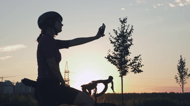 剪影的年轻女子站在旁边的自行车和自拍或照片在手机上日落。在金色的夕阳下，女自行车手站在城市建筑背景的山上。旅行的生活视频下载