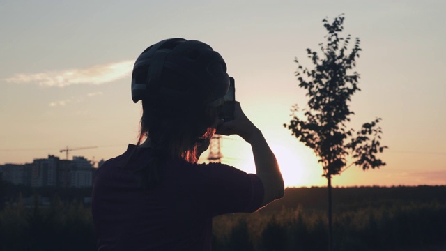 女孩正在用手机拍摄美丽的日落。一个骑自行车的女人的剪影正在用手机拍下阳光下的照片。女自行车旅行者站在旁边的自行车对金色的日落视频下载