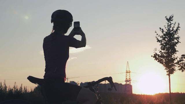 女游客在日落时分用手机拍照。女人骑着自行车站在公园里，对着美丽的阳光，用手机拍照。旅行的概念。戴着头盔的女孩剪影视频下载