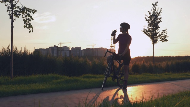 骑自行车的女人在夕阳下的公园里骑车后喝水。年轻的女运动员在自行车训练后喝水的肖像，站在自行车道的阳光下。铁人三项的概念视频下载