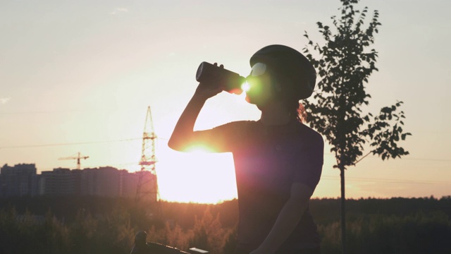 年轻美丽的女人在日落喝水的肖像。女自行车手正在喝水，经过艰苦的训练后，在充满阳光的公园里。自行车和铁人三项概念。健康的生活方式视频下载