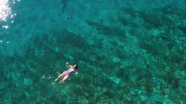 年轻的女性漂浮在背后的明星姿势和放松在温暖的绿松石亚得里亚海海浪与岩石海岸。悠闲的人度假时间概念空中俯瞰4K镜头。视频素材