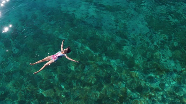 年轻的女性漂浮在背上，放松在温暖的碧绿的亚得里亚海海浪与岩石海岸。悠闲的人度假时间概念空中俯瞰4K镜头。视频素材