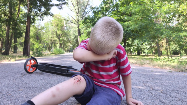 一个四岁的小孩从踏板车上摔下来，伤了膝盖。孩子哭着，揉着膝盖，朝它吹气。粗心驾驶和父母粗心大意的概念。视频下载