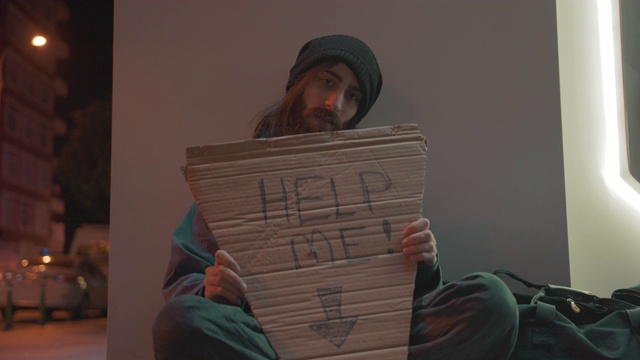 一个留着胡子和长发的无家可归的乞丐坐在街上寻求帮助的慢动作。拿着“帮我”的牌子。无家可归的概念,4 k。视频下载
