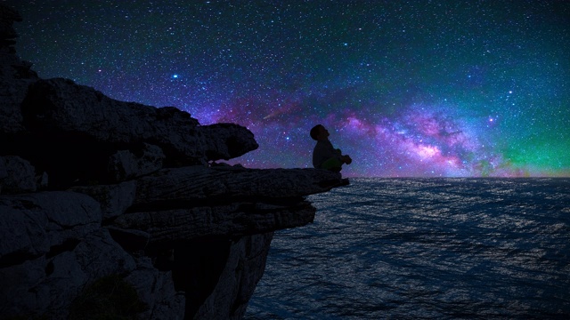 一个男孩的剪影坐在悬崖边看着银河和流星视频素材