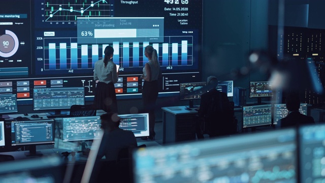 两名软件工程师在现代监控办公室的大屏幕旁交谈，实时分析馈电与图表。大数据科学家和管理者坐在电脑前。视频素材