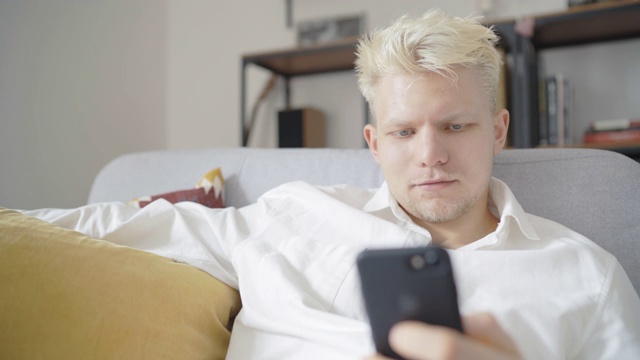 4K亮金发的男人坐在沙发上，手里拿着手机在浏览互联网视频素材
