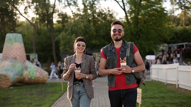 一对时髦的夫妇在城市公园散步吃着冰淇淋和华夫饼开怀大笑。浪漫的年轻男女在户外享受他们的闲暇时光。男朋友和女朋友的慢动作视频素材