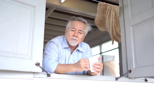 年长的亚洲男人看着窗边，拿着一杯茶在家视频素材