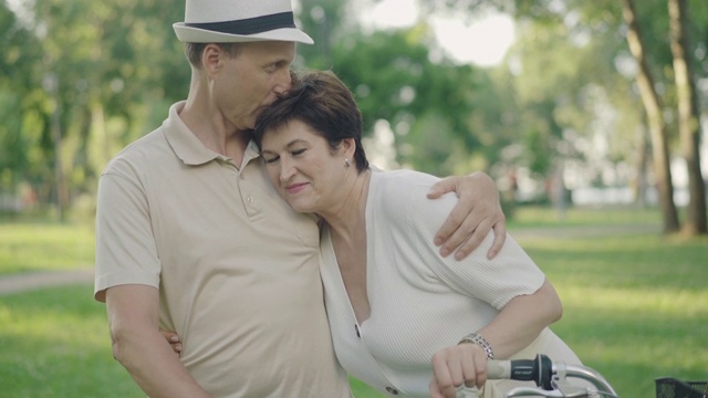 优雅的中年丈夫拥抱美丽的妻子并亲吻她的头。周末在阳光明媚的公园里休息的幸福的白人家庭的肖像。愉快的夫妇在户外享受夏日。视频下载