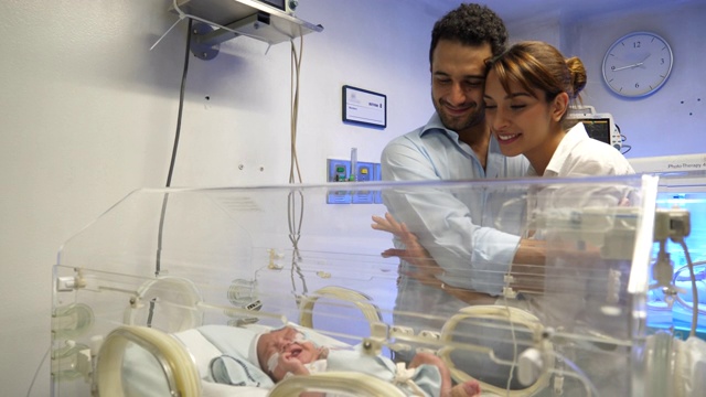 在新生儿重症监护室里，一对幸福的夫妇深情地看着他们在恒温箱里的新生儿视频素材