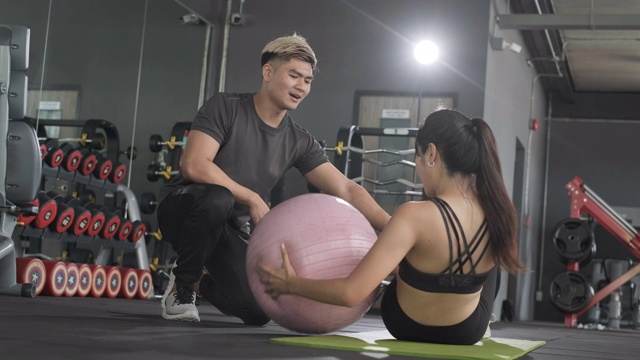 年轻的亚洲女子做俄罗斯旋转瑜伽球和锻炼与私人教练协助她和给予指导。女性健身与男性教练帮助照顾和教学视频下载