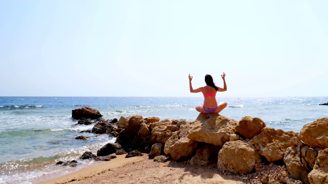 后视图。一个穿着泳衣的年轻女子坐在海边沙滩的石头上冥想，练习瑜伽。身体护理和冥想概念视频素材