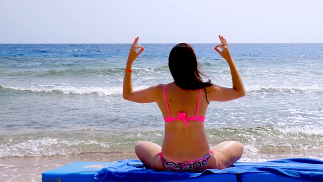 后视图。一个穿着泳衣的年轻女子坐在沙滩上的躺椅上冥想，练习瑜伽。身体护理和冥想概念视频素材