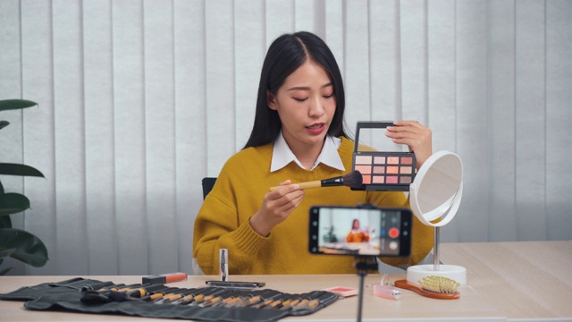 一位年轻的亚洲女性在家里用手机相机拍摄了一段关于化妆品的视频，并在她的博客上写了关于美容产品的视频。视频素材
