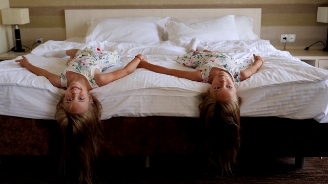 在一家旅馆的卧室里，两个双胞胎女孩倒立着躺在床上笑着。视频素材