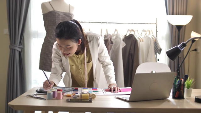 在服装店工作的女设计师使用笔记本电脑。时装设计师在纸上画出轮廓视频素材