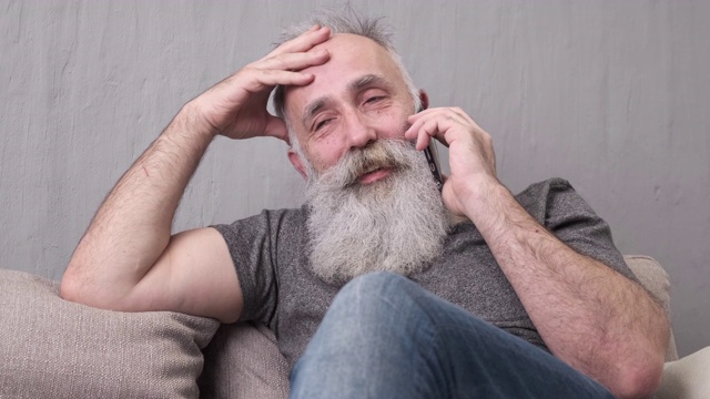留着胡子的老人坐在沙发上用智能手机聊天视频素材