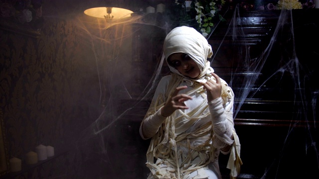 电影镜头的女孩穿着万圣节木乃伊服装摆姿势和吓人在旧的黑暗哥特式室内覆盖着蜘蛛网和雾与变化的照明。4K慢镜头50帧/秒视频下载
