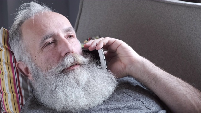 长着胡子的老人躺在沙发上用智能手机聊天视频素材