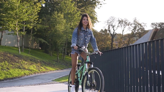 可爱的微笑辣妹在牛仔裤衣服骑自行车附近的金属栅栏视频素材